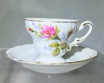 Può includere: Un set da tè bianco con tazza e piattino con un motivo floreale con rose rosa e foglie verdi. La tazza ha un bordo dorato e il piattino ha un bordo ondulato.