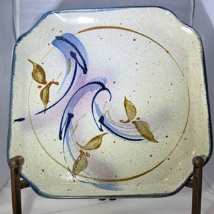 May include: A square ceramic plate with a cream background and a blue and brown abstract design. The design features swirling lines and leaves.