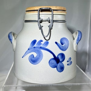 May include: A white ceramic jar with a blue floral design and a wooden lid with a metal latch. The jar has a traditional style and is perfect for storing dry goods.