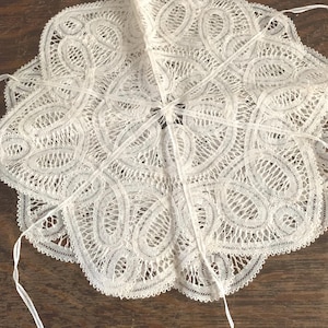 May include: A circular off-white lace doily featuring an intricate repeating paisley-inspired pattern with openwork mesh details and scalloped edges. The piece includes thin white fabric ties extending from the center and outer perimeter.