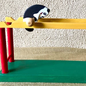 May include: A wooden toy featuring a black and white skunk figure on a yellow track. The toy has red support posts and a green base. The skunk has a detailed face and is positioned as if sliding down the track. The toy is painted with bright colors.