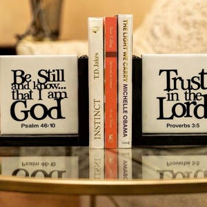 May include: White ceramic bookends with black text that reads "Be Still and know... that I am God" and "Trust in the Lord". The bookends hold several books with titles like "Instinct" and "The Light We Carry".