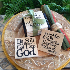 May include: Four beige square coasters with black lettering. The coasters feature religious quotes. The coasters are stacked and tied with a green ribbon. The coasters are on a wooden tray with a carved design. The tray is on a tiled surface.