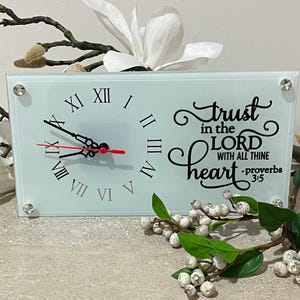 May include: A rectangular glass clock with Roman numerals and black hands, the second hand is red. The clock face has the words "Trust in the Lord with all thine heart" in black script. The clock is decorated with clear crystals.