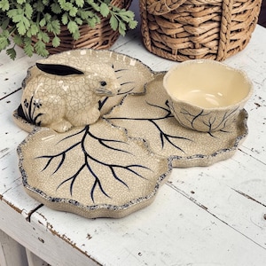 May include: A ceramic serving set featuring a leaf-shaped tray with a rabbit figurine and a small bowl. The set has a cream-colored base with dark blue leaf vein detailing and a crackle finish. The rabbit has black accents.