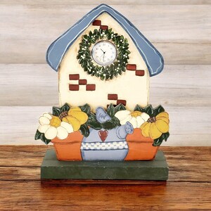 May include: A wooden house-shaped clock with a blue roof, white brick facade, and a wreath with a clock face. The house is decorated with red brick accents and a flower pot with sunflowers and daisies. The base is painted green.