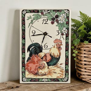 May include: A rectangular wall clock with a cream-coloured face featuring black numerals 3, 6, 9, and 12. The clock displays a colourful illustration of a rooster and a hen, surrounded by a decorative border of green leaves, white flowers, and dark berries.