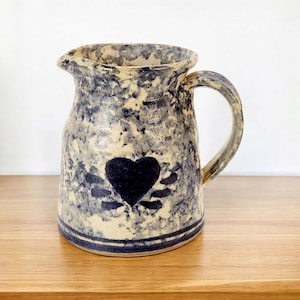 Puede incluir: Una jarra de cerámica azul y blanca con un diseño de corazón. La jarra tiene un esmalte moteado y un borde azul.