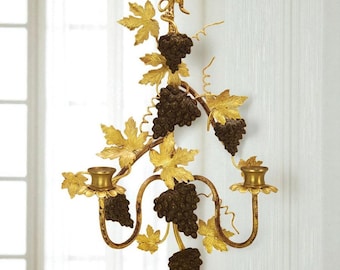 May include: A decorative wall sconce featuring a grape vine design. The sconce has two candle holders and is made of gold-colored metal with brown grape clusters and gold leaves. The sconce is mounted on a white wall.