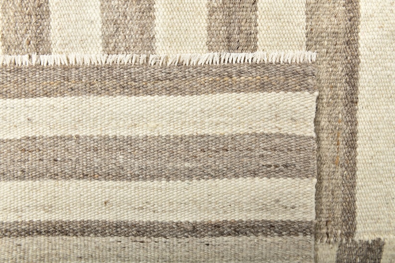 May include: A close-up of a woven rug with a striped pattern in shades of beige and brown. The rug has a fringed edge.