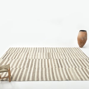 May include: A striped rug with a light brown and white pattern. A small wooden stool with a woven seat sits on the rug. A terracotta vase is in the background.