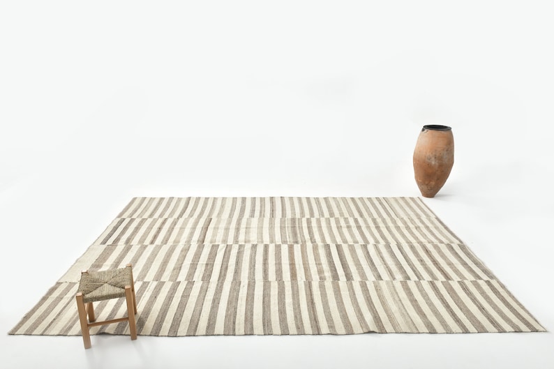 May include: A striped rug with a light brown and white pattern. A small wooden stool with a woven seat sits on the rug. A terracotta vase is in the background.