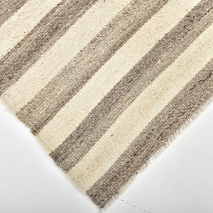 May include: Close-up of a woven rug with a striped pattern in shades of beige and gray. The rug has a frayed edge.