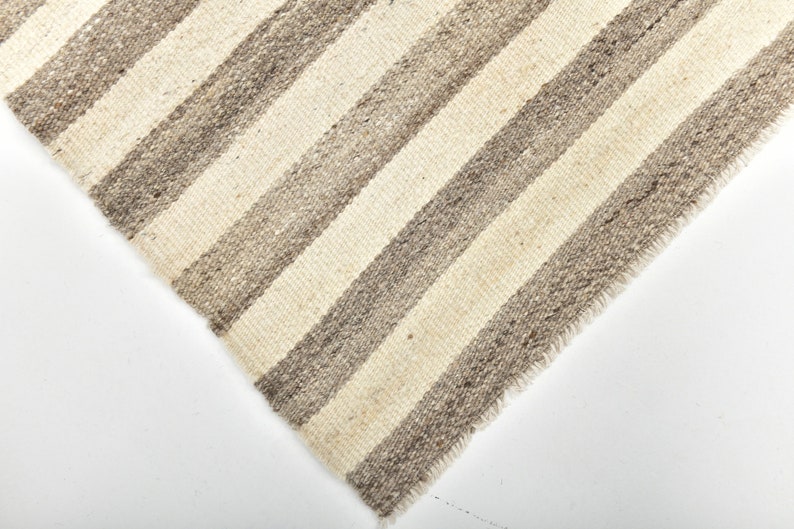 May include: Close-up of a woven rug with a striped pattern in shades of beige and gray. The rug has a frayed edge.