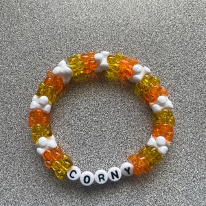Pode incluir: Uma pulseira de contas laranja, amarelas e brancas. A palavra "CORNY" é escrita com contas de letras brancas. As contas estão dispostas em um padrão circular, criando um acessório divertido e colorido.