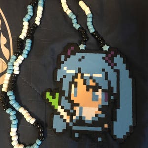 May include: A beaded necklace featuring a pixelated anime character pendant. The pendant is primarily blue, black, and white, with a green element. The necklace has a beaded chain in black, white, and light blue.