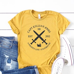 Könnte beinhalten: Ein senfgelbes T-Shirt mit einem schwarzen Grafikdesign mit gekreuzten Schwertern, einem Schwan und dem Text "CAPT KILLIAN JONES EST SB 2012 A PIRATE'S LIFE-IT'S FOREVER".