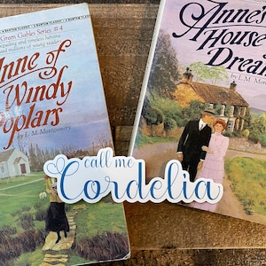May include: Two paperback books by L.M. Montgomery, "Anne of Windy Poplars" and "Anne's House of Dreams", from the Anne of Green Gables series. A blue and white sticker with the text "call me Cordelia" is placed on the front cover of the first book.