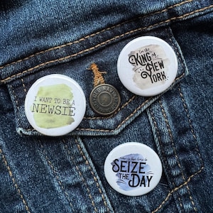 Puede incluir: Tres botones redondos con diferentes diseños. El primer botón es verde y blanco con el texto "Quiero ser un newsie". El segundo botón es blanco con un fondo de acuarela y el texto "Soy el rey de Nueva York". El tercer botón es azul y blanco con el texto "Ahora es el momento de aprovechar el día".