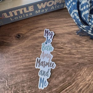 May include: A sticker with the text "Meg Jo Beth Amy Marmee and Me" in a vertical arrangement. The sticker has a white border and a gradient of colors, including blue, pink, and green. A copy of "Little Women" by Louisa May Alcott is in the background.