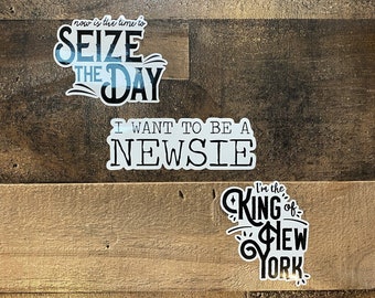 May include: Three Newsies-themed stickers on a wood background. The first sticker says "now is the time to SEIZE THE DAY". The second sticker says "I WANT TO BE A NEWSIE". The third sticker says "I'm the KING of NEW YORK".