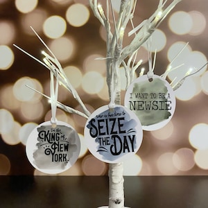 May include: Three round ornaments with watercolor backgrounds and black text. The first ornament says "I'm the King of New York". The second ornament says "Now is the time to Seize the Day". The third ornament says "I want to be a Newsie".