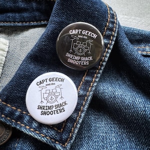 May include: Two round buttons with the text "CAPT GEECH and the SHRIMP SHACK SHOOTERS" in a stylized font. One button is black with white text, and the other is white with black text. The buttons are on a denim jacket.