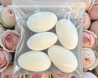 May include: A transparent plastic bag holding five white, oval sweets. The sweets are smooth and opaque. The background is filled with numerous pink and cream-coloured artificial roses.