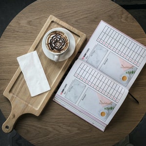 May include: A wooden cutting board with a white napkin, a cup of coffee with chocolate designs, and a pink and white planner with the text "Busy Bee Planner" and "Daily Planner".