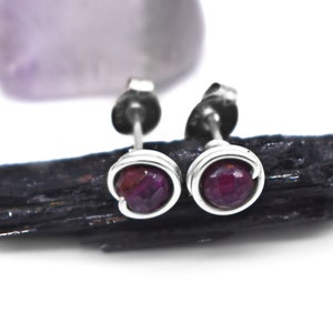 May include: Silver stud earrings featuring a deep red gemstone, set within a silver wire frame. The earrings are presented on a black, textured surface, with a blurred, pale purple stone in the background. The earrings are on a white background.