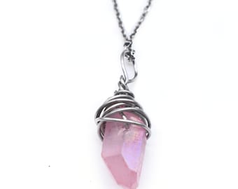 May include: A necklace featuring a raw, translucent pink crystal point pendant wrapped in dark gray metal wire. The pendant hangs from a matching dark gray metal chain, showcasing a faceted, iridescent surface that reflects light with subtle hues.