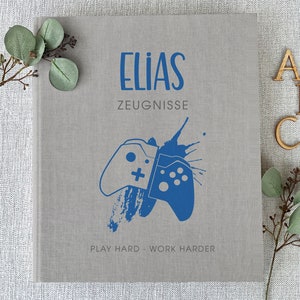 Könnte beinhalten: Ein graues, mit Leinen bezogenes Tagebuch mit dem Namen "ELIAS" in blauer Schrift. Das Tagebuch hat eine blaue Grafik mit zwei Videospielcontrollern und dem Text "ZEUGNISSE" und "PLAY HARD - WORK HARDER" unterhalb der Grafik.