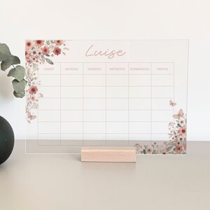 Könnte beinhalten: Ein transparenter Acryl-Wochenplaner mit floralem Design und dem Namen "Luise" oben. Der Planer hat Spalten für jeden Tag der Woche, wobei die Tage auf Deutsch beschriftet sind.