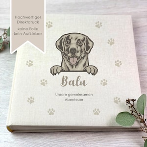 Könnte beinhalten: Ein beiges Fotoalbum mit einem braunen Hund, der über die Oberseite schaut. Der Hund hat eine braune Nase und die Zunge hängt heraus. Das Album hat den Text "Balu" und "Unsere gemeinsamen Abenteuer" auf der Vorderseite.