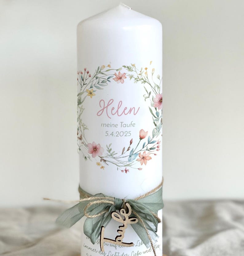 May include: A white pillar candle decorated with a floral wreath and the name "Helen" in pink script. The candle is adorned with a green ribbon, twine, and a wooden charm. The date "5.4.2025" is also printed on the candle.