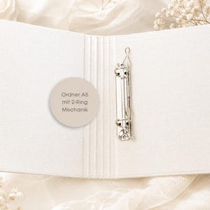May include: An open A5 binder with a white fabric cover and a silver metal 2-ring mechanism. A circular beige sticker is attached to the inside cover, featuring the text: Ordner A5 mit 2-Ring Mechanik. The binder has a textured, woven appearance.