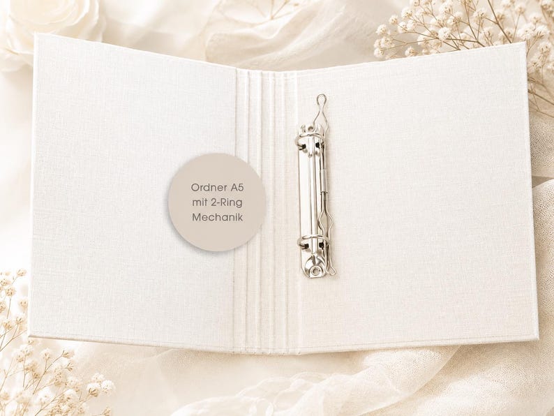 May include: An open A5 binder with a white fabric cover and a silver metal 2-ring mechanism. A circular beige sticker is attached to the inside cover, featuring the text: Ordner A5 mit 2-Ring Mechanik. The binder has a textured, woven appearance.