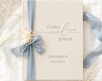 May include: A cream-coloured fabric-covered book titled Stammbuch with the names Caro und Jonas and the date 2016 1 25 printed on the front. The book is decorated with a dusty blue frayed ribbon tied in a bow and a small cluster of dried white flowers.