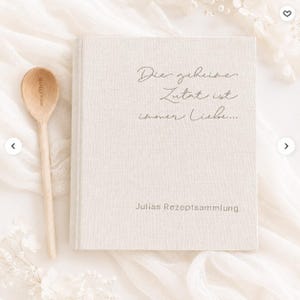 May include: A beige linen-covered recipe book with the German text "Die geheime Zutat ist immer Liebe..." and "Julias Rezeptsammlung" printed on the front cover, accompanied by a light-colored wooden spoon with the engraved text "MARLEY SPOON".