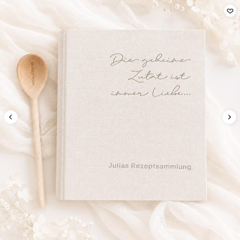 May include: A beige linen-covered recipe book with the German text "Die geheime Zutat ist immer Liebe..." and "Julias Rezeptsammlung" printed on the front cover, accompanied by a light-colored wooden spoon with the engraved text "MARLEY SPOON".