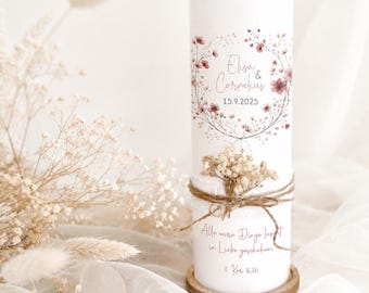 Puede incluir: Una vela cilíndrica blanca con una ilustración de corona floral en tonos rosa suave, los nombres Elisa & Cornelius y la fecha 15.9.2025. Está envuelta con cuerda y flores secas, incluyendo el texto Alle eure Dinge lasst in Liebe geschehen.