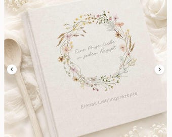 May include: A square, off-white fabric-covered recipe book featuring a watercolor floral wreath illustration. The text reads "Eine Prise Liebe in jedem Rezept" inside the wreath and "Elenas Lieblingsrezepte" at the bottom. A wooden spoon is visible.