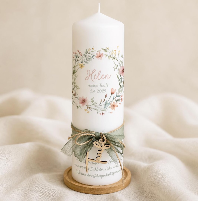 May include: A white pillar candle decorated with a floral wreath and the name "Helen" and date "5.4.2025". The candle is adorned with a green ribbon, wooden charm, and sits on a wooden base. The candle is a decorative item.