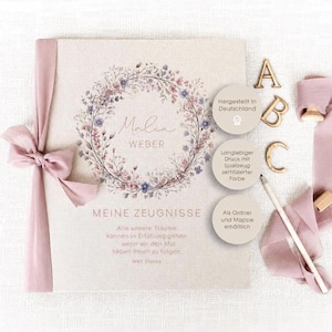 Könnte beinhalten: Ein personalisiertes Erinnerungsbuch mit Leinenstruktur, verziert mit einem floralen Kranz und dem Namen "Malia Weber". Der Text "Meine Zeugnisse" ist sichtbar. Ein rosa Band und Holzbuchstaben "A, B, C" sind ebenfalls vorhanden.