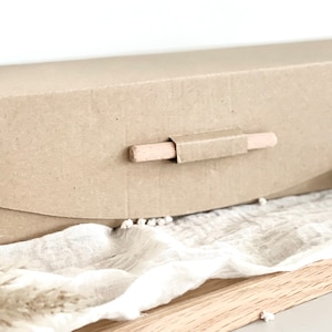 May include: A rectangular, light brown cardboard box with a wooden handle. The box is closed and has a curved flap on the front. The handle is made of a small wooden dowel. The box is sitting on a white cloth.