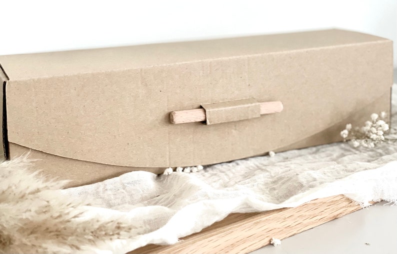 May include: A rectangular, light brown cardboard box with a wooden handle. The box is closed and has a curved flap on the front. The handle is made of a small wooden dowel. The box is sitting on a white cloth.