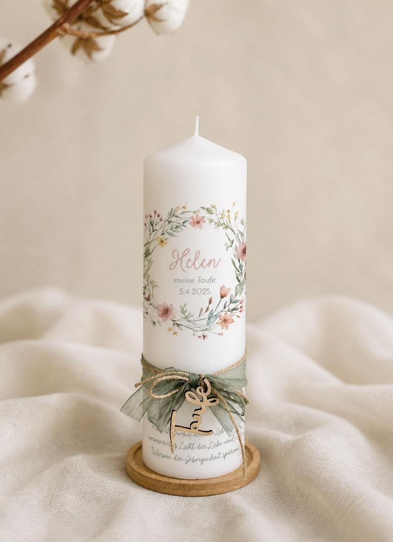 May include: A white pillar candle decorated with a floral wreath, the name "Helen" and the date "5.4.2025". The candle is adorned with a green ribbon and a wooden tag. The candle sits on a small wooden base.