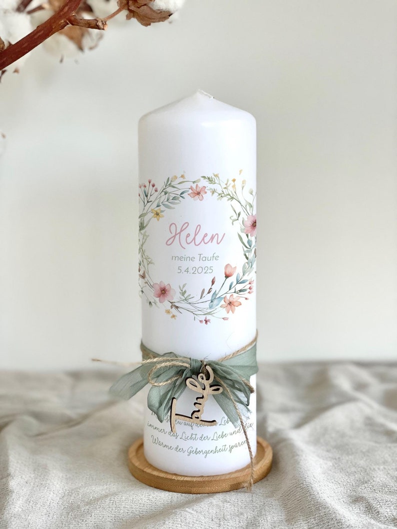 May include: A white pillar candle decorated with a floral wreath and the name "Helen". The candle is adorned with a green ribbon, a wooden charm, and rests on a wooden coaster. The text "meine Taufe 5.4.2025" is also visible.