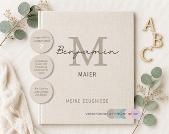 Può includere: Un raccoglitore quadrato rivestito in tessuto beige chiaro con il nome Benjamin Maier, una grande lettera M e il testo MEINE ZEUGNISSE. Tre badge circolari mostrano testo in tedesco e le lettere in legno A, B e C sono posizionate a destra.