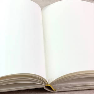 May include: A blank, white book with a linen cover and a yellow ribbon bookmark. The book is open and lying flat on a light wood surface.